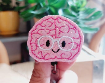 Op de afbeelding: Een roze stoffen brein met een witte omtrek en glitter, met een witte bril en een glimlach. Het brein is bevestigd aan een zwarte intrekbare badgehouder.