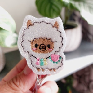 Könnte beinhalten: Ein weißer Glitzer-Lama mit braunen Augen und einer rosa Blume, der eine bunte Perlenkette mit Quasten trägt. Der Lama ist an einem durchsichtigen Plastik-Badgehalter befestigt.