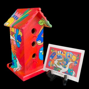 Puede incluir: Una colorida casita de pájaros de madera con una base roja y un techo multicolor. La casita tiene tres agujeros para que los pájaros puedan entrar. Una pequeña tarjeta con una ilustración colorida y el texto "La Lutte Portrait de Famille Enfance" se muestra en un soporte negro delante de la casita de pájaros.