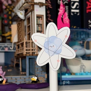 May include: A white sticker with a cartoon atom design. The atom has a blue and white nucleus with a smiling face and pink hearts around it.