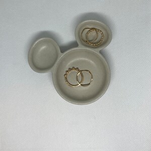 May include: A gray ceramic ring dish shaped like Mickey Mouse with two gold rings inside.