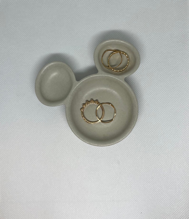 May include: A gray ceramic ring dish shaped like Mickey Mouse with two gold rings inside.