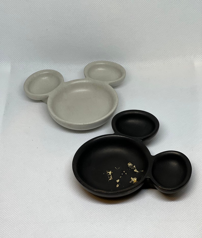 May include: Two ceramic Mickey Mouse shaped dishes. One dish is gray and the other is black. The black dish has gold flakes inside.