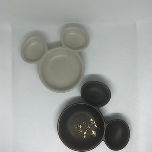 May include: Two ceramic Mickey Mouse shaped bowls. One bowl is beige and the other is black with gold flecks.