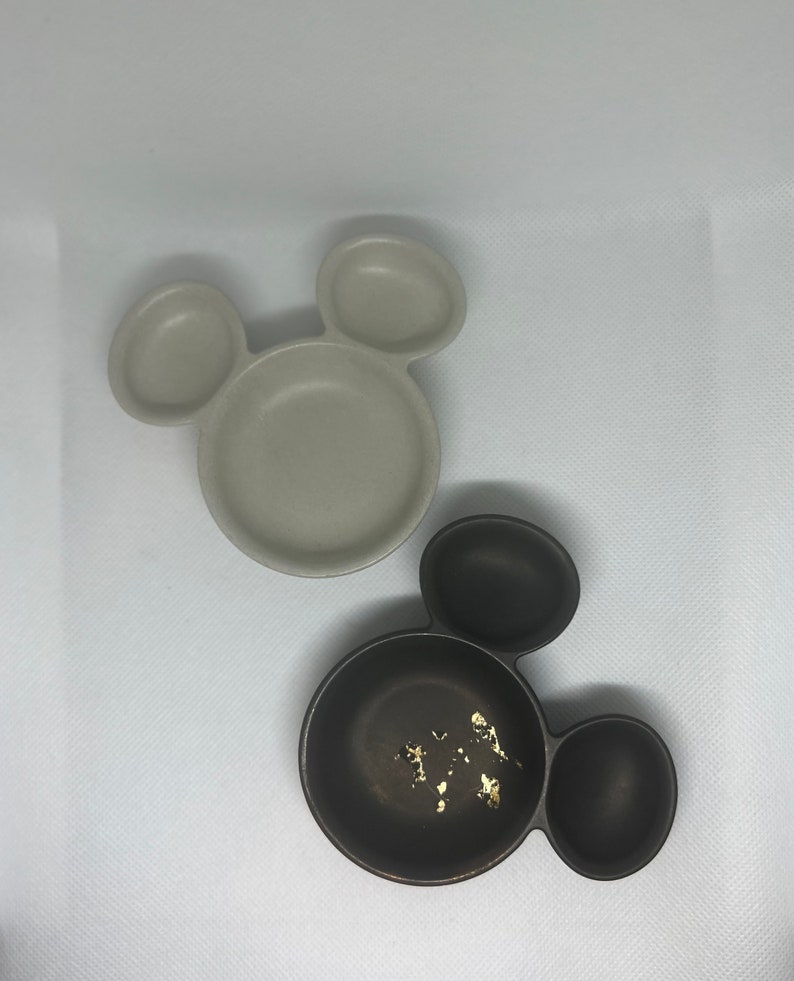 May include: Two ceramic Mickey Mouse shaped bowls. One bowl is beige and the other is black with gold flecks.