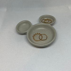 May include: A gray ceramic ring dish shaped like Mickey Mouse with two gold rings inside.