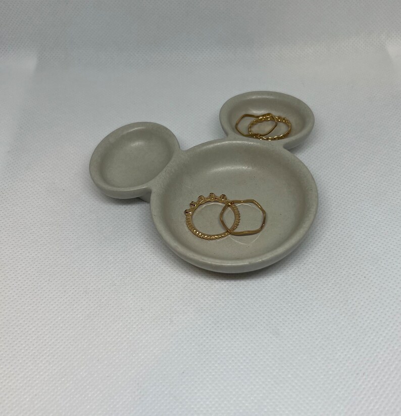 May include: A gray ceramic ring dish shaped like Mickey Mouse with two gold rings inside.