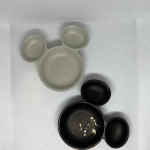 May include: Two Mickey Mouse shaped ceramic dishes. One is gray and the other is black with gold flecks.