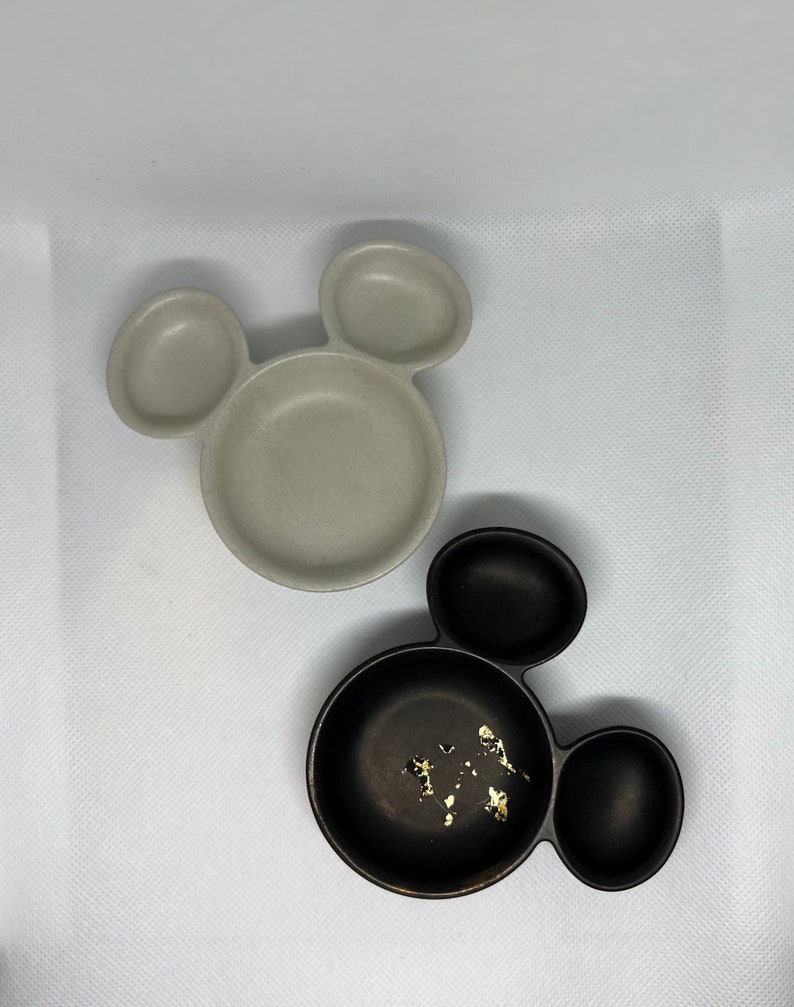May include: Two Mickey Mouse shaped ceramic dishes. One is gray and the other is black with gold flecks.