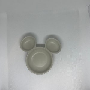 May include: A light gray ceramic dish shaped like Mickey Mouse with three sections. The dish is perfect for serving snacks or dips.