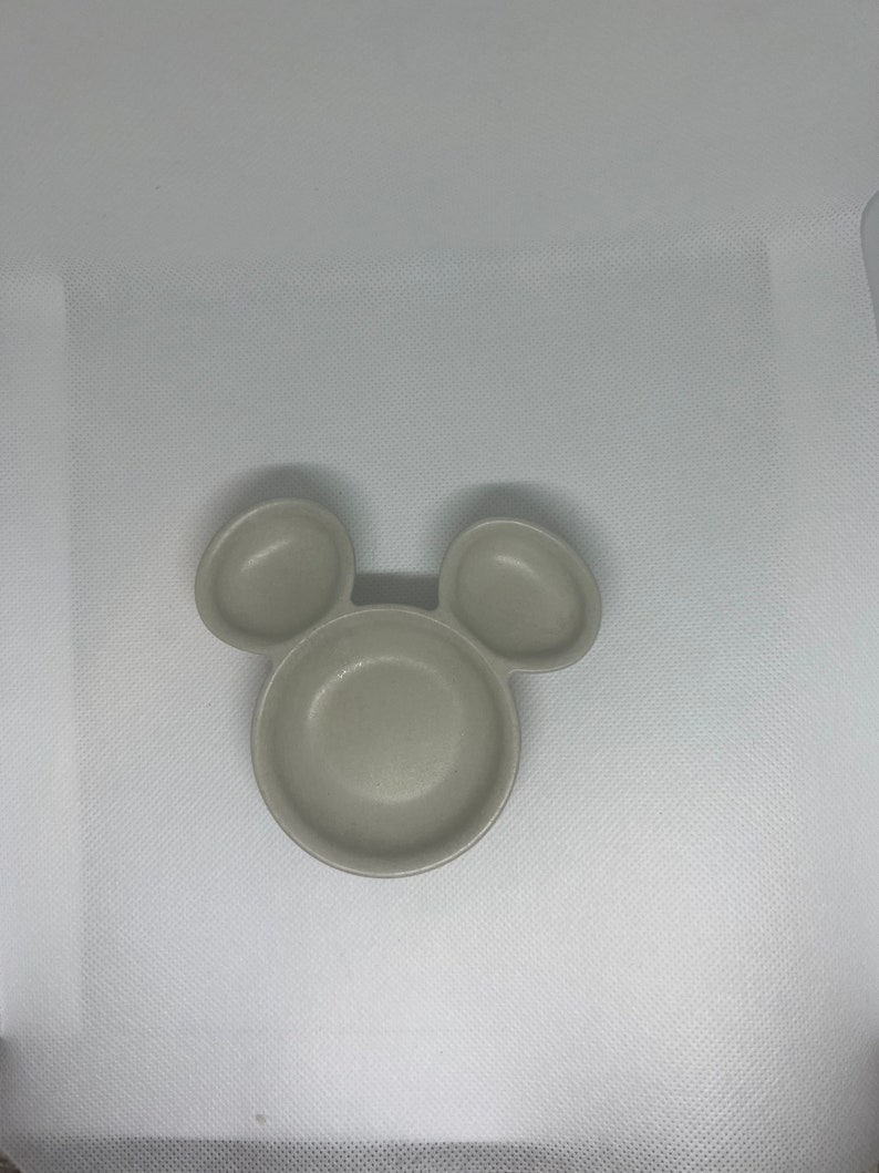 May include: A light gray ceramic dish shaped like Mickey Mouse with three sections. The dish is perfect for serving snacks or dips.