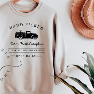 May include: A beige sweatshirt with a black graphic design of a pickup truck carrying pumpkins. The text reads "HAND PICKED Farm Fresh Pumpkins PUMPKINS GOURDS APPLES - OPEN DAILY - "