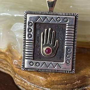 May include: Silver pendant with a gold bail and a square frame. The pendant features a hand design with a red gemstone in the center. The frame has geometric patterns and a dark background.