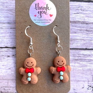 May include: Gingerbread man earrings with red bow ties and white buttons. The earrings are on a brown card with a thank you sticker that says "Thank you for supporting my small business".