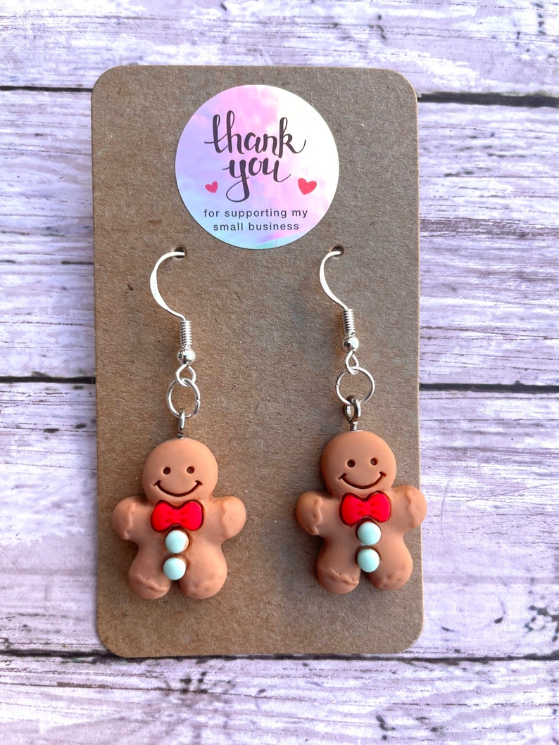 May include: Gingerbread man earrings with red bow ties and white buttons. The earrings are on a brown card with a thank you sticker that says "Thank you for supporting my small business".