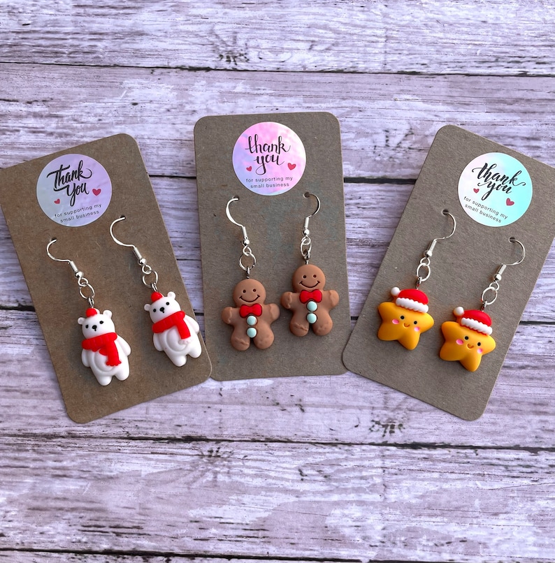 May include: Three pairs of Christmas-themed earrings displayed on brown card stock. The earrings are silver with a white polar bear, a gingerbread man, and a yellow star with a red Santa hat. Each card has a round sticker with the text "Thank you for supporting my small business".