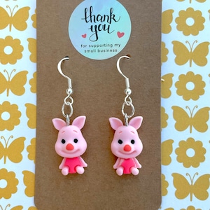 May include: A pair of piglet earrings with silver hooks. The piglets are pink with black eyes and snouts. The earrings are on a brown card with a thank you sticker that says "Thank you for supporting my small business".