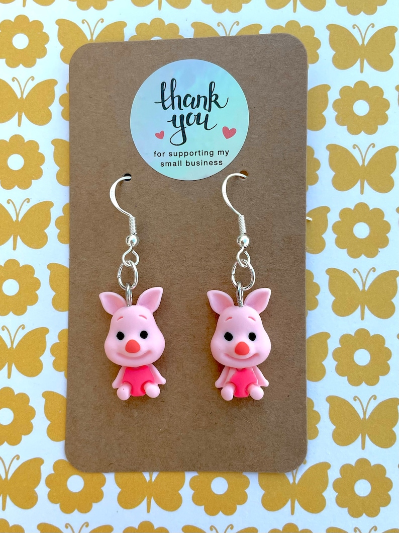 May include: A pair of piglet earrings with silver hooks. The piglets are pink with black eyes and snouts. The earrings are on a brown card with a thank you sticker that says "Thank you for supporting my small business".