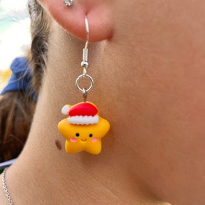 May include: A pair of silver dangle earrings with a yellow star charm wearing a red Santa hat. The star has a smiling face.