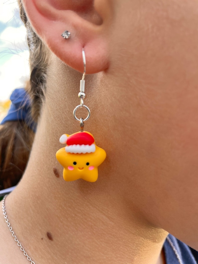 May include: A pair of silver dangle earrings with a yellow star charm wearing a red Santa hat. The star has a smiling face.
