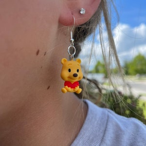 May include: A pair of dangle earrings featuring a yellow Winnie the Pooh character charm. The charm is wearing a red shirt and has a brown nose.