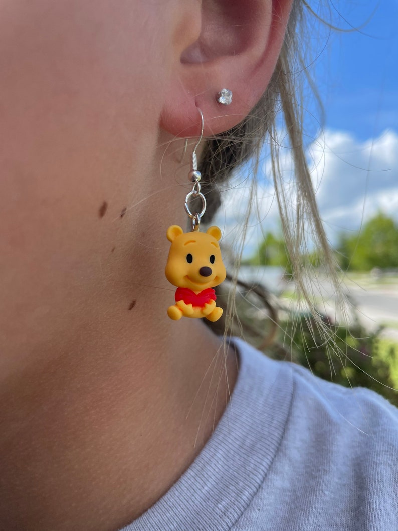 May include: A pair of dangle earrings featuring a yellow Winnie the Pooh character charm. The charm is wearing a red shirt and has a brown nose.