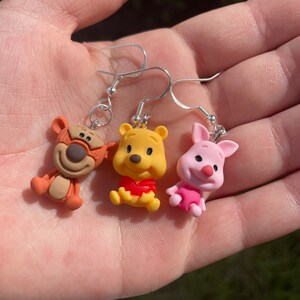 May include: Three Winnie the Pooh character earrings. The earrings are silver with brown, yellow, and pink clay charms of Tigger, Winnie the Pooh, and Piglet.