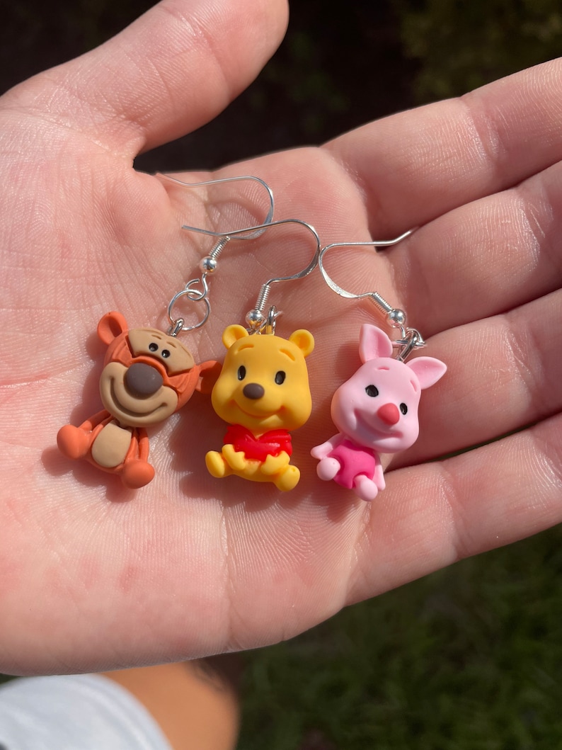 May include: Three Winnie the Pooh character earrings. The earrings are silver with brown, yellow, and pink clay charms of Tigger, Winnie the Pooh, and Piglet.