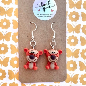 May include: A pair of dangle earrings featuring a cartoon orange and brown character with a black face. The earrings are on a brown card with a white circle sticker that says "thank you" with two red hearts and the text "for supporting my small business".