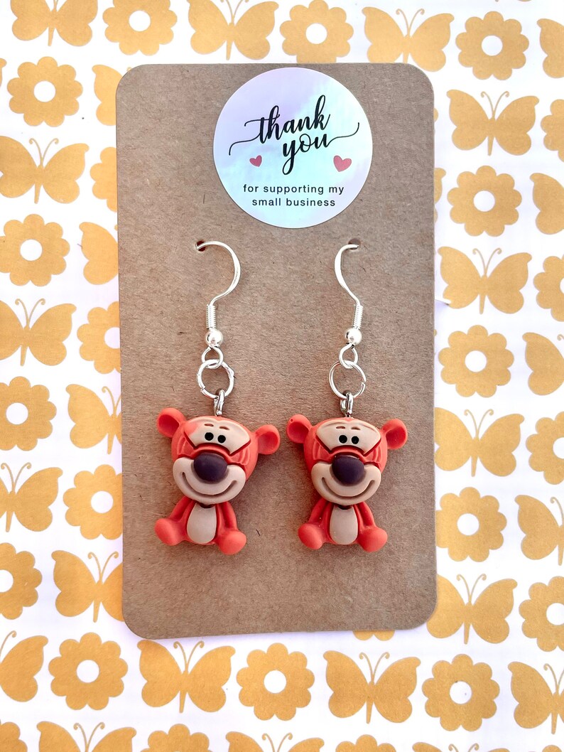 May include: A pair of dangle earrings featuring a cartoon orange and brown character with a black face. The earrings are on a brown card with a white circle sticker that says "thank you" with two red hearts and the text "for supporting my small business".