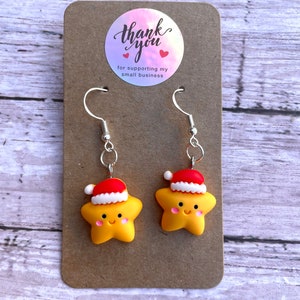 May include: A pair of yellow star-shaped earrings with red and white Santa hats. The earrings are on a brown card with a thank you sticker that says "Thank you for supporting my small business".