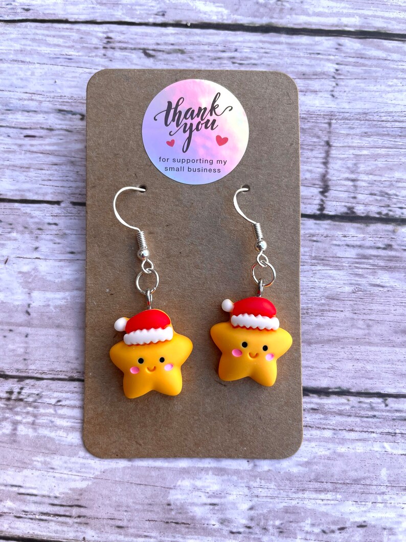 May include: A pair of yellow star-shaped earrings with red and white Santa hats. The earrings are on a brown card with a thank you sticker that says "Thank you for supporting my small business".