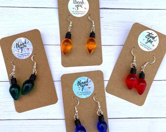 May include: Four pairs of dangle earrings featuring lightbulb charms. The charms are in red, blue, green, and orange. Each pair is displayed on a brown card with a thank you sticker.