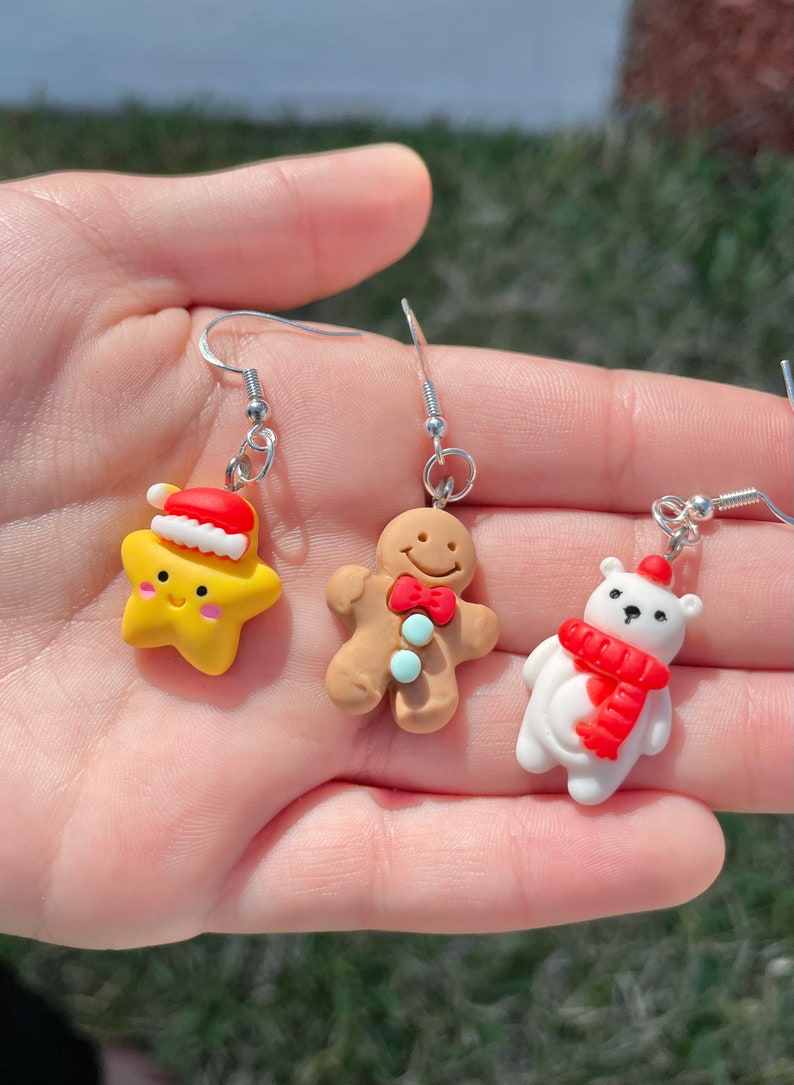 May include: Three pairs of Christmas-themed earrings featuring a gingerbread man, a star with a Santa hat, and a polar bear wearing a red scarf.