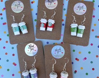 May include: Five pairs of novelty earrings, each designed as a miniature coffee cup in shades of teal, red, green, purple, and pink. The earrings are displayed on brown cards with a sticker that reads "Thank you for supporting my small business."