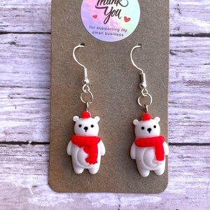 May include: A pair of silver dangle earrings featuring white polar bear charms with red hats and scarves. The earrings are displayed on a brown card with a sticker that says "Thank You for supporting my small business".