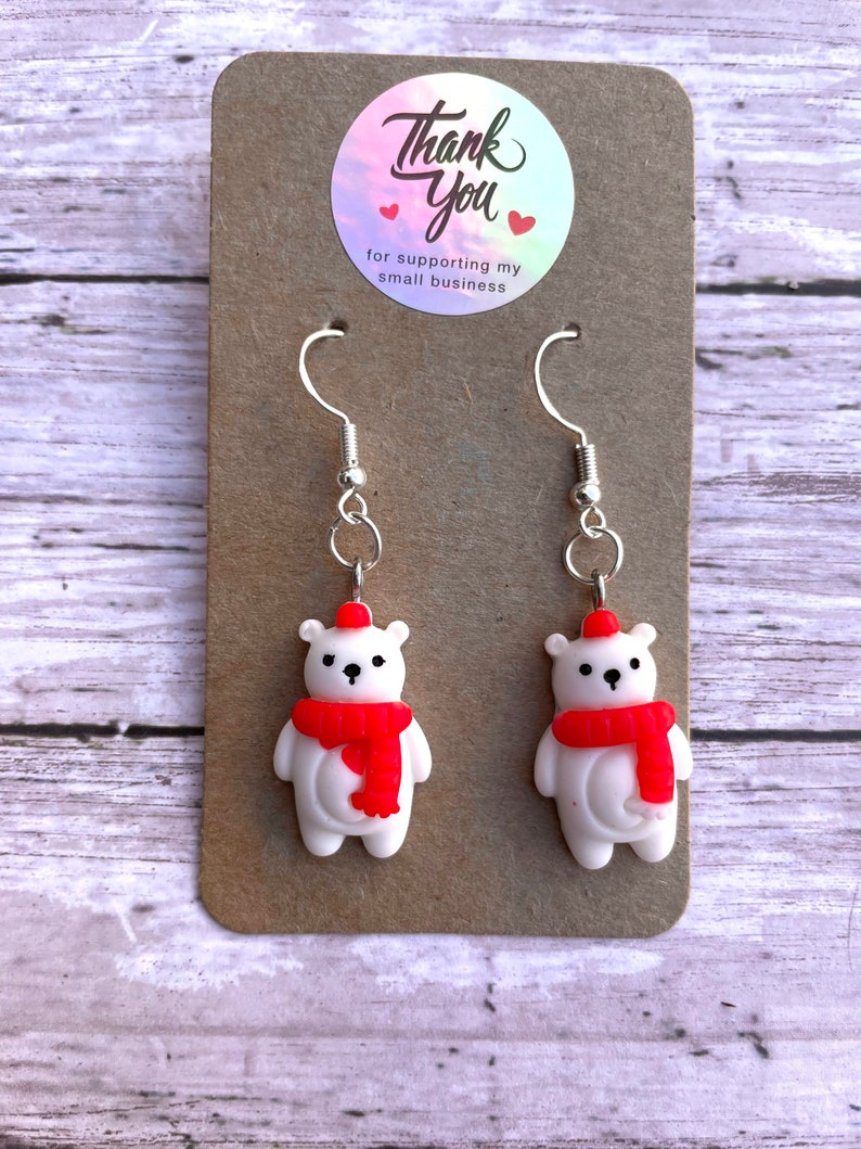 May include: A pair of silver dangle earrings featuring white polar bear charms with red hats and scarves. The earrings are displayed on a brown card with a sticker that says "Thank You for supporting my small business".