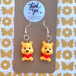 May include: A pair of Winnie the Pooh dangle earrings with silver hooks. The earrings feature a yellow cartoon Winnie the Pooh character with a red shirt. The earrings are displayed on a brown card with a thank you sticker.