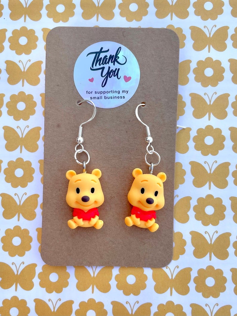 May include: A pair of Winnie the Pooh dangle earrings with silver hooks. The earrings feature a yellow cartoon Winnie the Pooh character with a red shirt. The earrings are displayed on a brown card with a thank you sticker.