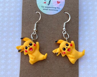 May include: A pair of yellow Pikachu earrings with black-tipped ears and red cheeks, suspended from silver-coloured hooks. The earrings are presented on a brown card with a holographic sticker that reads "thank you for supporting my small business".