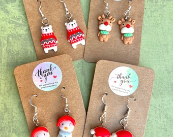 May include: Four pairs of Christmas-themed dangle earrings. The earrings are made of polymer clay and feature a snowman, Santa Claus, a polar bear wearing a red sweater, and a reindeer. Each pair of earrings is displayed on a brown card with a thank you message for supporting small businesses.