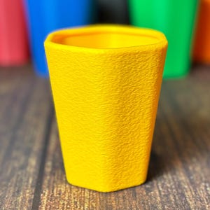 May include: A yellow, textured cup is in focus, with a blurred background of red, blue, green, and orange cups. The cups have a geometric design and are arranged on a wooden surface. The cups are likely for beverages.