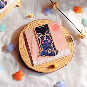 Può includere: Un personaggio dei cartoni animati nero e blu con un naso rosa e un anello giallo intorno al collo. Il personaggio tiene una versione più piccola di se stesso. Il personaggio è su uno sfondo rosa e bianco con stelle e una luna. Il testo "#morbidart" è nell'angolo in basso a destra dell'immagine.