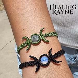 May include: Two macrame bracelets, one green and one black, each featuring a central gemstone and a crescent moon design. The bracelets are worn on a wrist. The text "HEALING RAYNE" is in the upper right corner.
