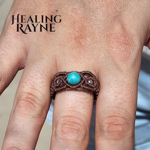 May include: A brown macrame ring featuring a central turquoise bead and smaller silver-colored beads. The ring is handmade with intricate knotting. The text "HEALING RAYNE" is in the upper left corner.