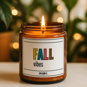 May include: A lit candle in an amber glass jar with a white label. The label features the text FALL in multicolored block letters, the word vibes in black, and the brand name CoNo Candle Co. at the bottom, framed by two horizontal black lines.