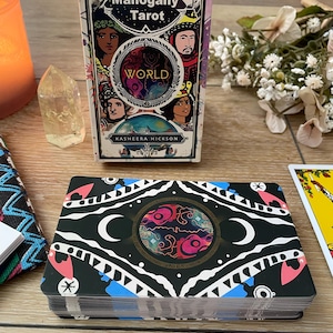 May include: A deck of Mahogany Tarot cards with a matching box. The box features the words "Mahogany Tarot" and "World" with illustrations of diverse faces. The card backs are black with a circular design, moons, and geometric shapes.