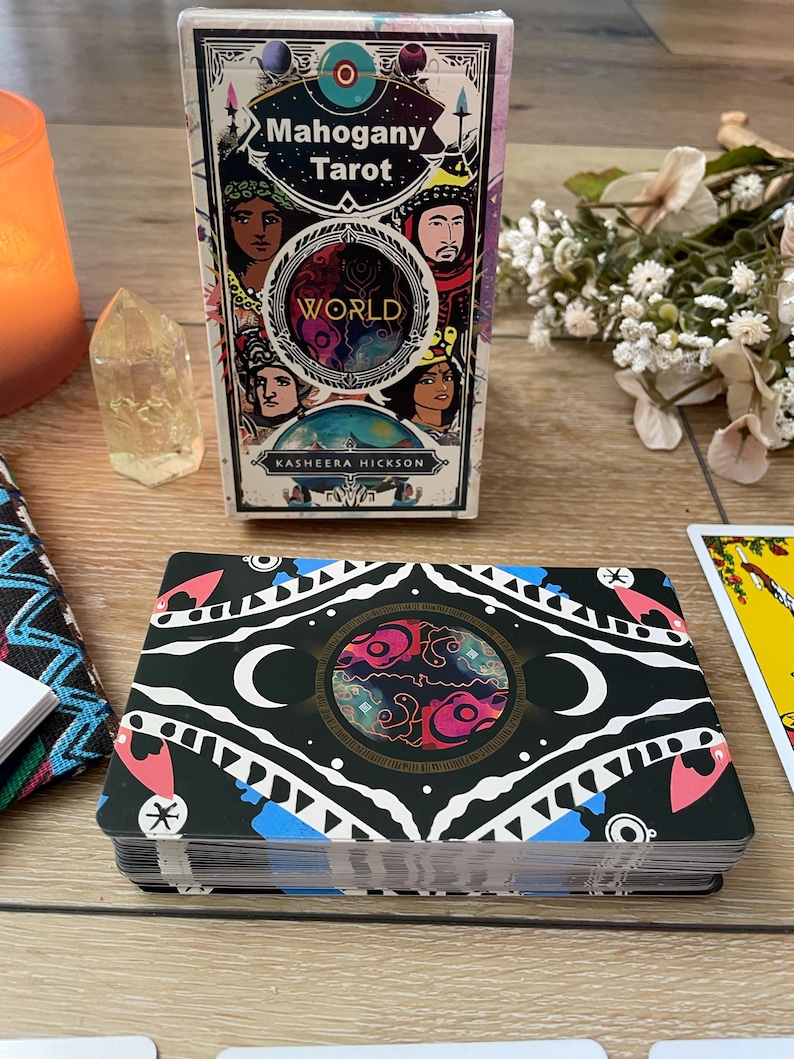 May include: A deck of Mahogany Tarot cards with a matching box. The box features the words "Mahogany Tarot" and "World" with illustrations of diverse faces. The card backs are black with a circular design, moons, and geometric shapes.