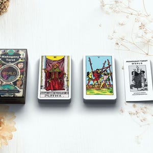 May include: An overhead shot of a tarot card set with a box and three card stacks. The box reads "Mahogany Tarot World" with illustrations. The cards show various tarot imagery, including "Justice" and "World". The set is on a white surface.