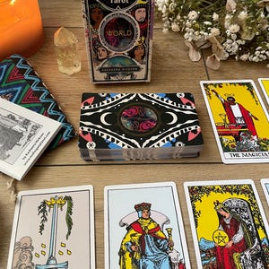 May include: A deck of "Mahogany Tarot" cards with a decorative box featuring diverse faces and the word "WORLD". Several tarot cards are displayed, along with a candle, crystal, and small bouquet. The cards have colorful illustrations.